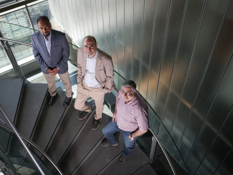 Three men stand on a spiral staircase looking up at the camera.