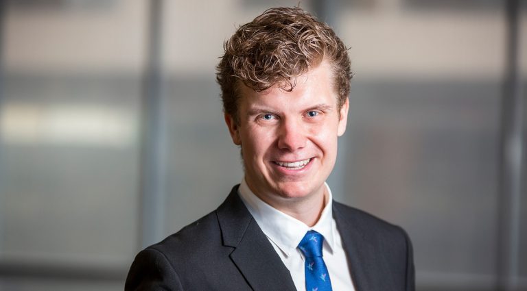 Smiling young man with short, curly blonde hair, blue eyes and wearing a white suit shirt, dark grey blazer and blue tie.