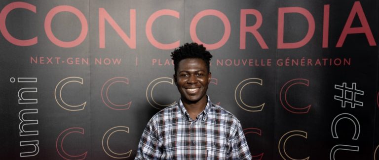 A young, smiling man with short, dark, spiky hair, wearing a plaid shirt and with a poster in the background with big letters saying, "Concordia."