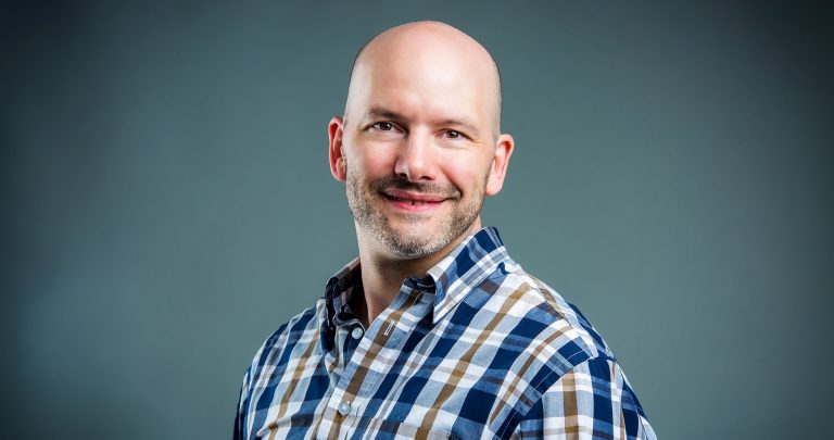 Smiling bald man wearing a checkered shirt.