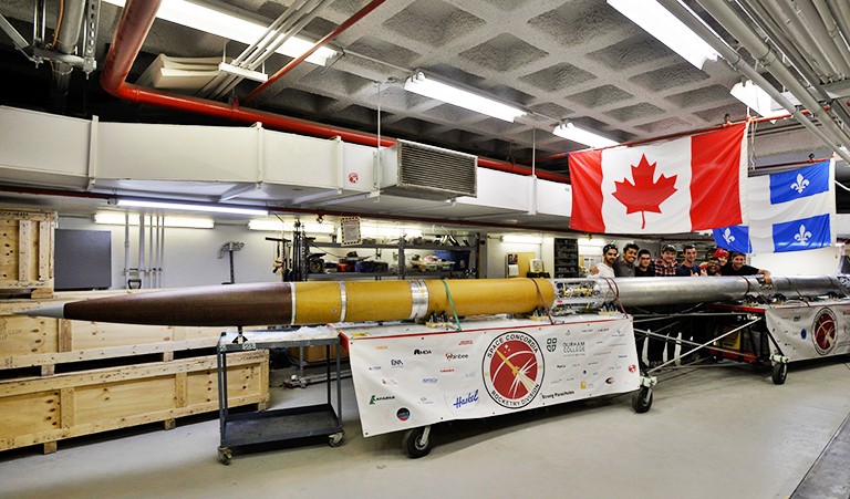A rocket lying on a trolley in a garage area with a team of young people near its base and a Canada and Quebec flag hung above.