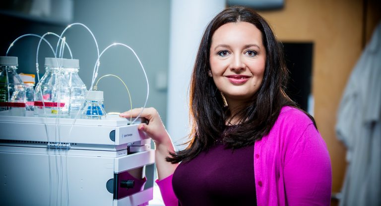 Smiling woman with long dark hair, wearing a burgundy top and a pink cardigan, resting her hand on a piece of machinery in a lab setting.