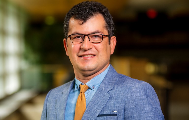 Smiling man with glasses and short, dark hair, wearing a blue dress shirt and blazer and a mustard-yellow tie.