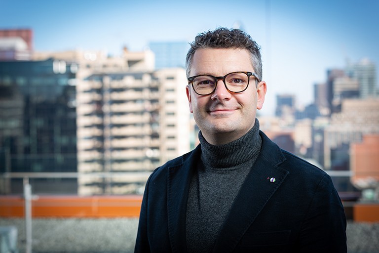 A smiling man with short, greying hair and glasses, wearing a grey polo neck sweater and a black blazer.