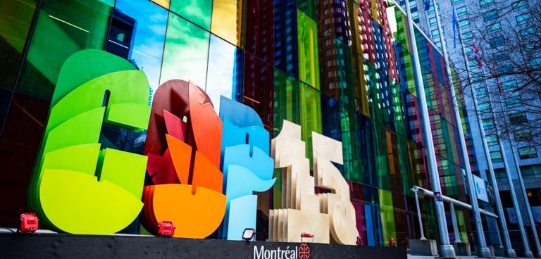 Giant green, red, blue and beige letter and numbers spell out the word COP15, with glass walled city buildings in the background.