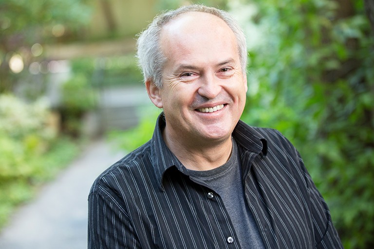 Smiling, balding man standing in an outdoor setting and wear a black lined open shirt over a grey T-Shirt.