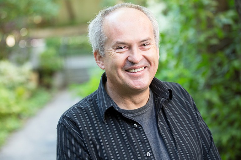 Concordia English professor and 2013 Man Booker International Prize finalist Josip Novakovich Smiling, balding man standing in an outdoor setting and wear a black lined open shirt over a grey T-Shirt.
