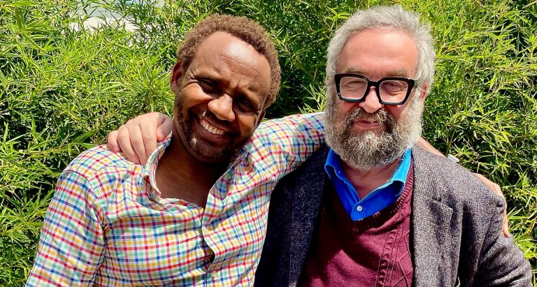 Two men standing in front of trees and with their arms around each other in a friendly embrace as they smile for the camera.