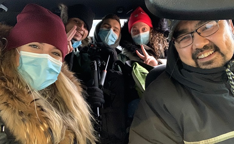 A group of people sitting in a car and wearing winter jackets with surgical masks on.