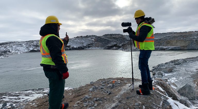 Two men in cold-weather gear, one with a camera, both wearing hardhats and boots and reflective vests, standing on a large rock with some patches of snow and a lake in the background.