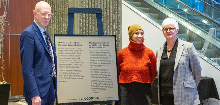 Three people — one man and two women — smiling for the camera and standing beside a large plaque.