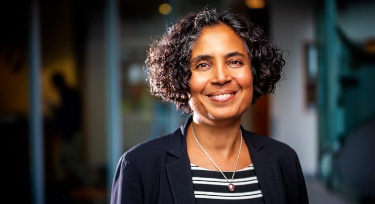 Smiling woman with shoulder-length curly hair, wearing a gold necklace, striped top and black blazer.