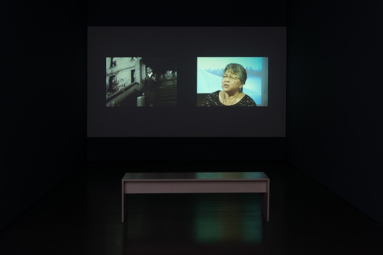 A screen in front of a desk with two images pictured, an archive image of a woman on stair, and an older woman with glasses speaking to someone off camera