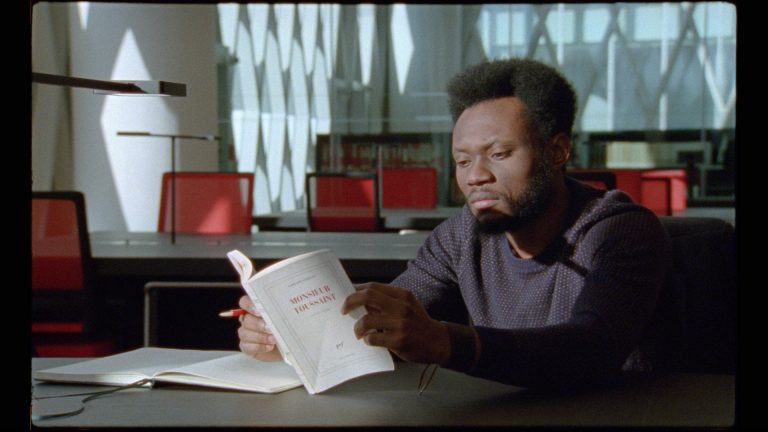 A man sitting at a desk in a library setting and reading a paperback book.