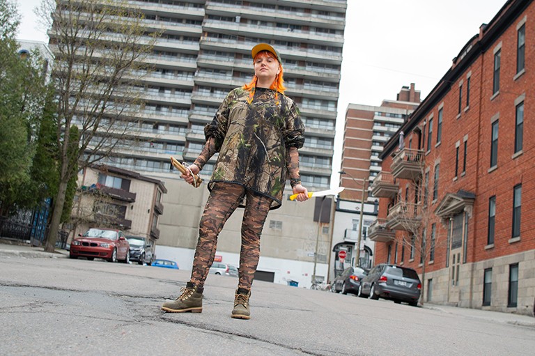 A person with long bright orange hair, wearing a cap and camouflage-style top and leggings, standing on an empty street and holding a knife and nunchucks. 