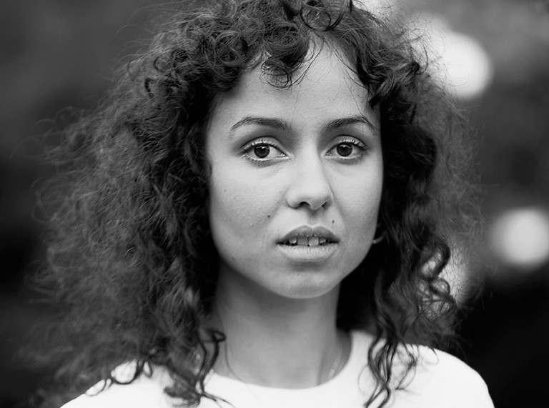 Black and white image of a woman with long, dark curly hair, wearing a white T-shirt and staring straight at the camera and 