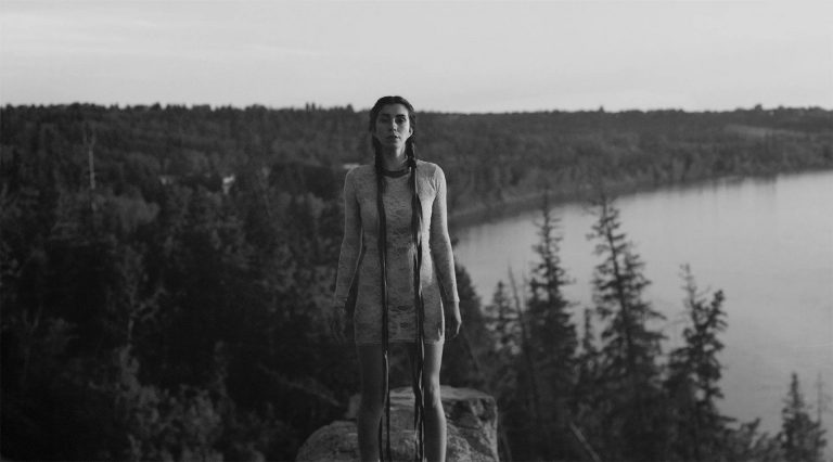 A woman in a fitting, semi-transparent lace dress with two long plaits down her front, standing on rock with a lake and forest far below her in the background.