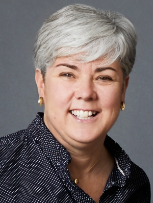 Smiling woman with short grey hair, wearing gold earrings, a gold necklace and a black shirt with small white polkadots