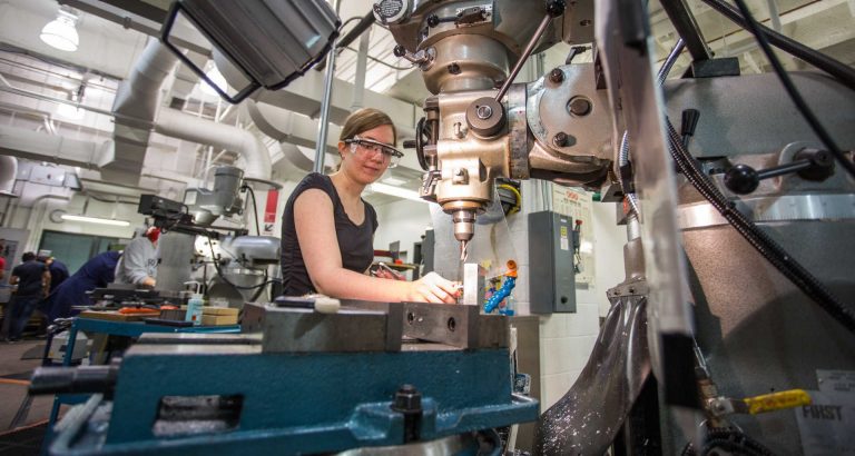 A young woman wearing a sleeveless top and safety goggles, in a university lab with large mechanical equipment.