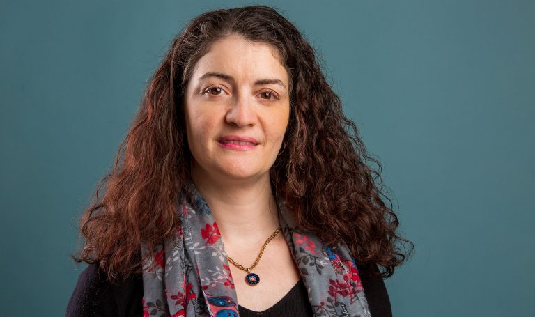 Slightly smiling woman with long, brown, curly hair, wearing a necklace, a scarf with grey with red patterns and a black top