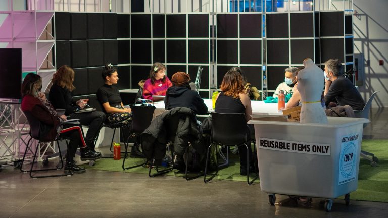 A group of young people sitting around a long table in an open space. In the foreground there is a trolley on wheels with a mannequin inside, and a sticker on the front saying, "Reusable items only."