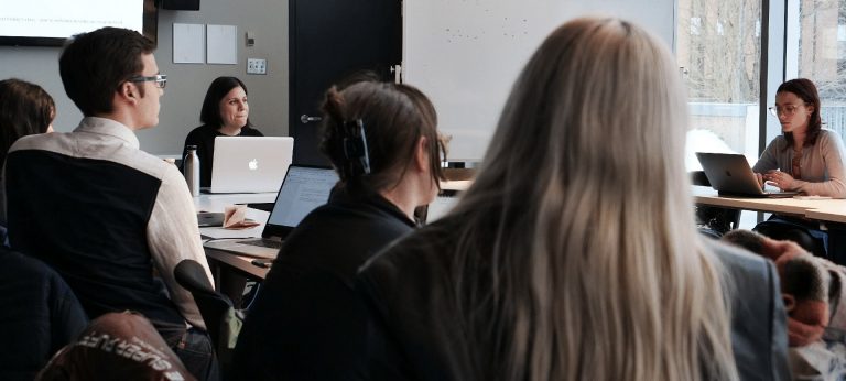 A classroom setting with students seated at two long desks and two women up front with laptops.