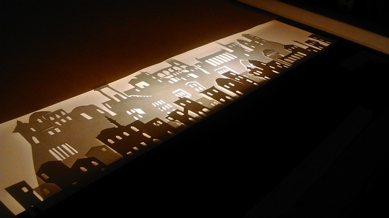 Soulikias assembles papercut silhouettes for a scene in his animated film "In the Shadow of Mount Damavand." An image in dark browns and sepia yellows of papercut silhouettes.