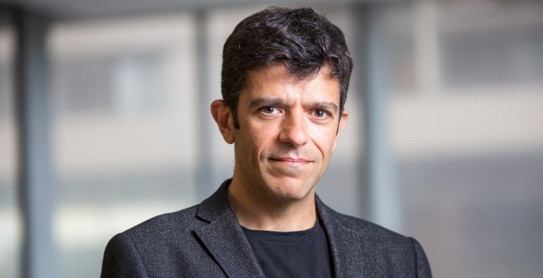 A slightly smiling man with short, dark hair and brown eyes, wearing a black T-Shirt and a dark grey blazer.