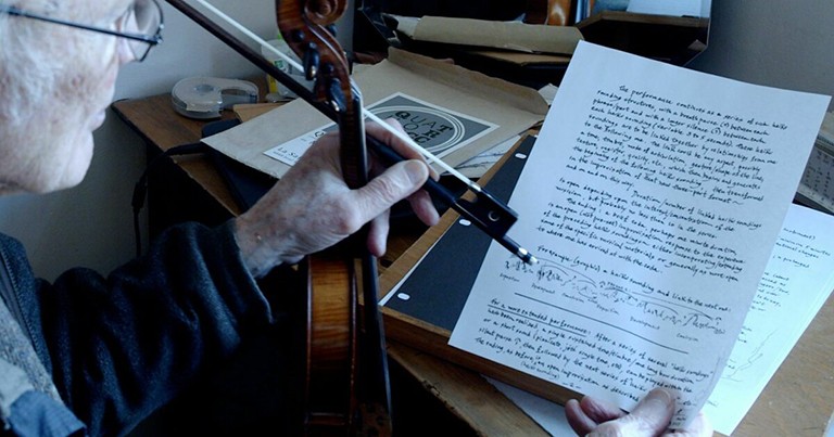 Photo taken from over the shoulder of an older man holding in one had a violin and bow and in the other a white piece of paper with black writing on it.