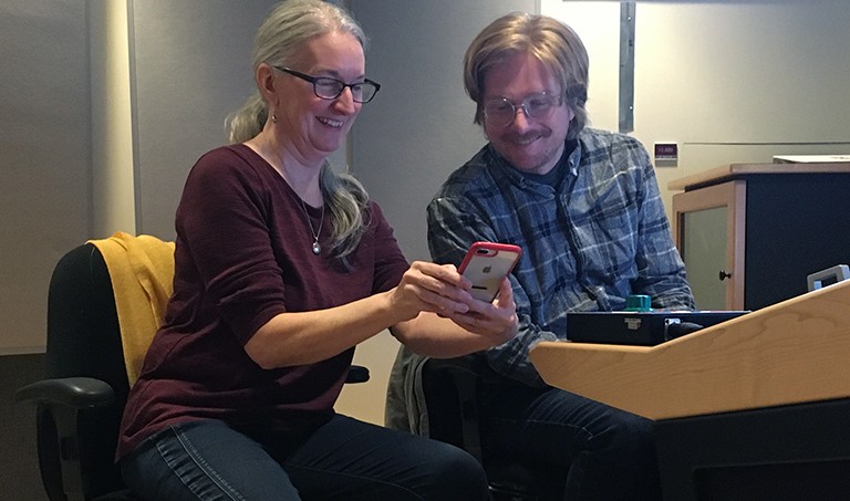 From left: Marion Mulvenna and Patrick McMaster in the recording studio. A smiling woman and man, both with glasses, looking at a cellphone that the woman is holding.