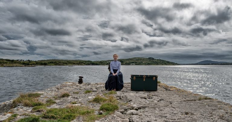 A woman in a buttoned up shirt and long skirt, sitting on the rocky shore of a wide lake.