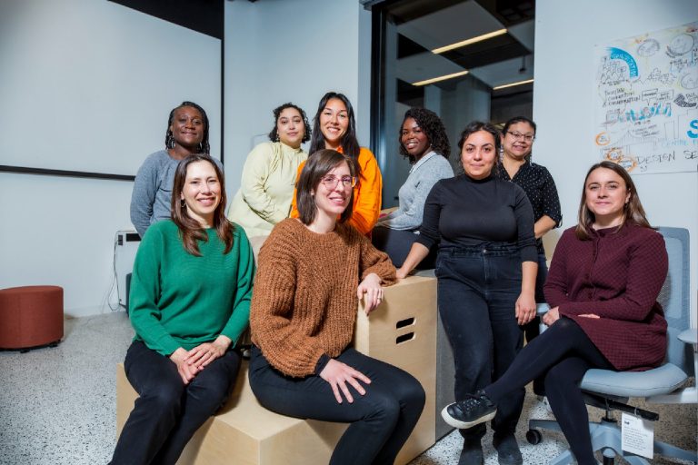 A group of women sitting and standing in a room, smiling at the camera.