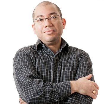 Smiling man with very close cropped hair and glasses, wearing a dark, checkered shirt.