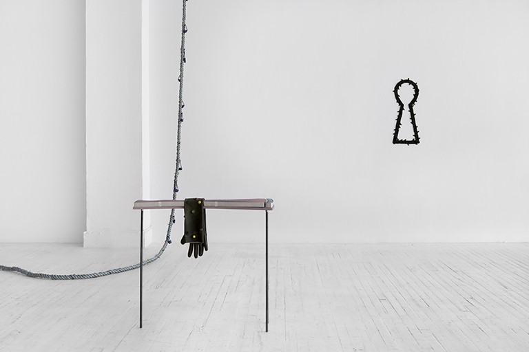 A white gallery space, with a rope snaking in from the left and a stand with one empty glove hanging on it, while a large sculpture like a keyhole hangs on the wall.