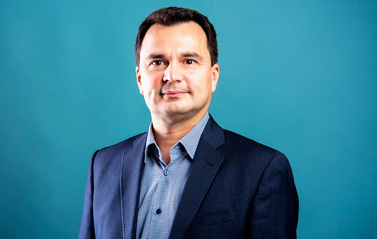 Slightly smiling man with short, dark hair combed back and wearing a light blue dress shirt with a dark blue blazer. 