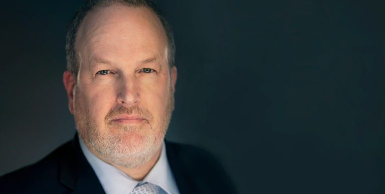 Close-up of a slightly smiling man with grey-blonde hair and beard, and wearing a suit and tie.