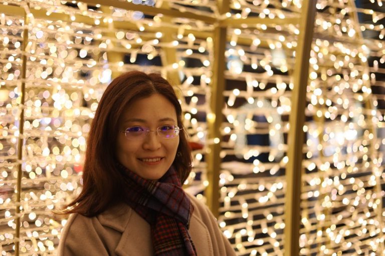 A women with glasses smiles. In the background blurred lights illuminate an outdoor night setting.