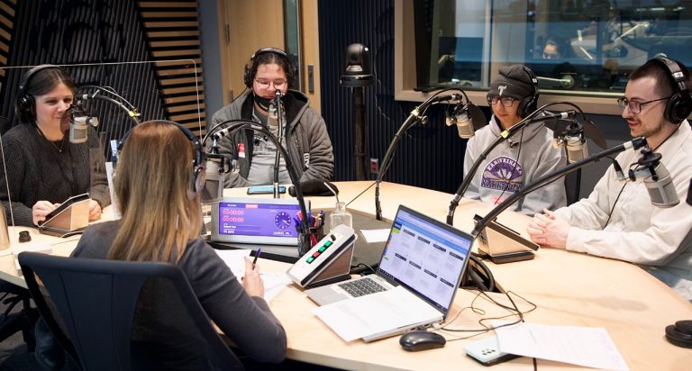 A group of young people with headphones on, sitting round a desk in a radio station studio.