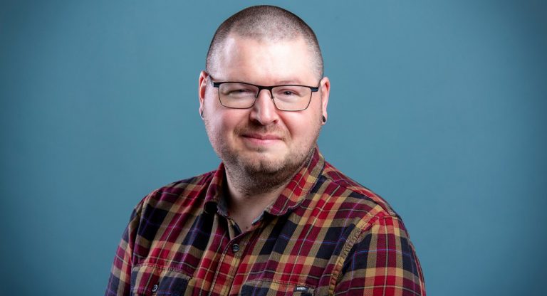 A smiling person with close-cropped dark hair and wearing glasses and a red, yellow and dark purple plaid shirt.