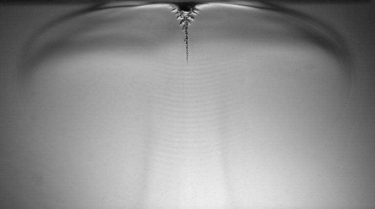 Image in black and white of what looks like sound waves making patterns in a white milk- or paint-like substance.