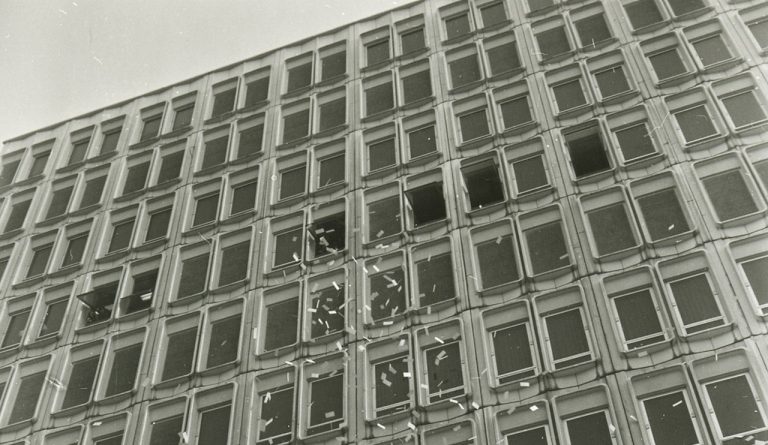 Black and white photo taken looking up at a city building with small pieces of paper fluttering down from the upper windows.