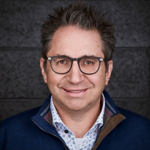 Photo portrait of a smiling man with dark hair and glasses wearing zip-up sweater with dress shirt underneath