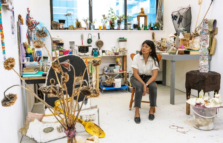 A woman with long dark hair sitting on a wooden chair in an art studio full of sculptures of found objects.