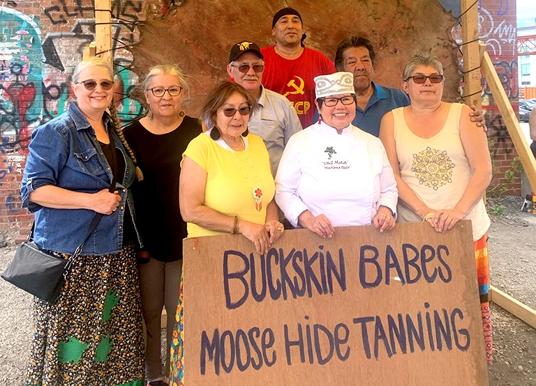 A group of men and women standing in an exterior location behind a sign that reads, "Buckskin Babes Moose Hide Tanning"