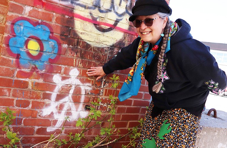 A woman in a hat, sunglasses and a long, black coat, standing beside a brick wall and putting her hand on the graffiti of the wall.