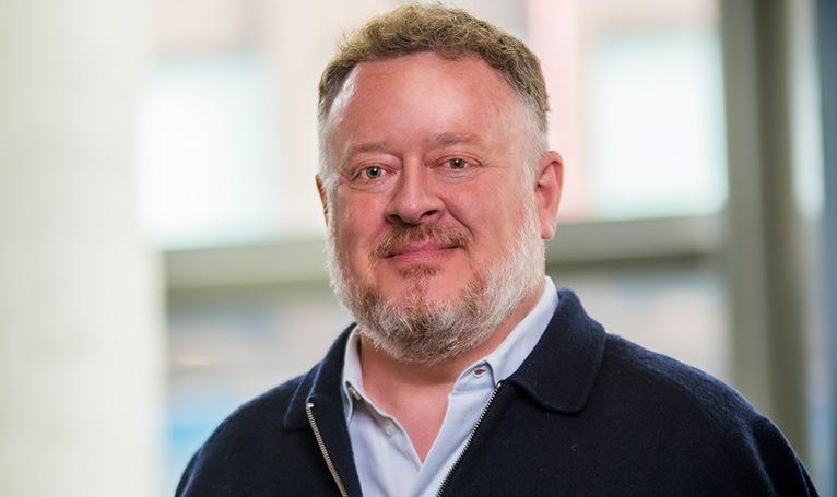 Smiling man with short blonde hair and a beard, wearing a light blue shirt and a zippered sweater.