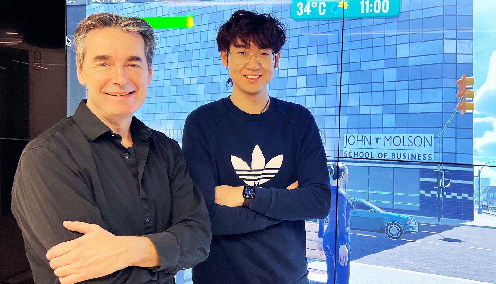 Christopher Gibbs, pictured with Jiarui Li, says of CityPlayer: “It’s Sim City with real science.” Two men standing together in front of a large screen with a video game on it, one older and one younger, smiling for the camera.