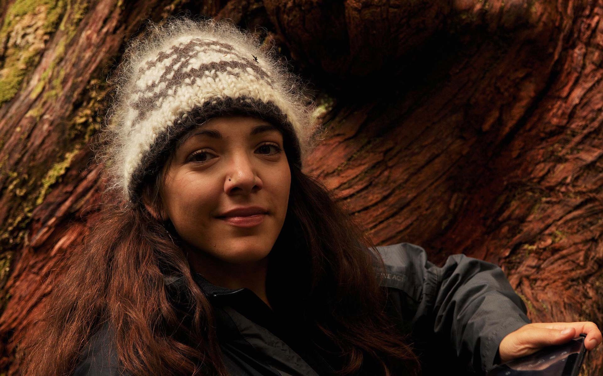 Sarah Moritz, a sociocultural anthropologist and Banting Postdoctoral Fellow. A smiling young woman in front on the trunk of an enormous tree, wearing a woollen beanie and a black jacket