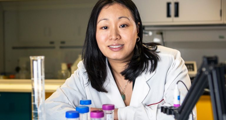 Smiling woman with long, dark hair in a lab coat sitting at a desk in a lab interior.