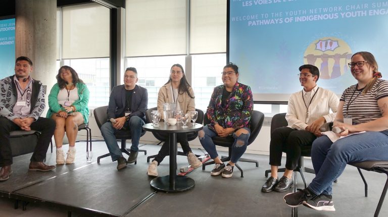 A group of young Indigenous youth leaders sit together on a stage.